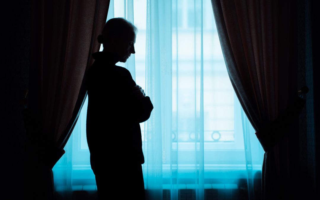 A man stands infront of a window with his head down, looking depressed. he may be suffering domestic abuse