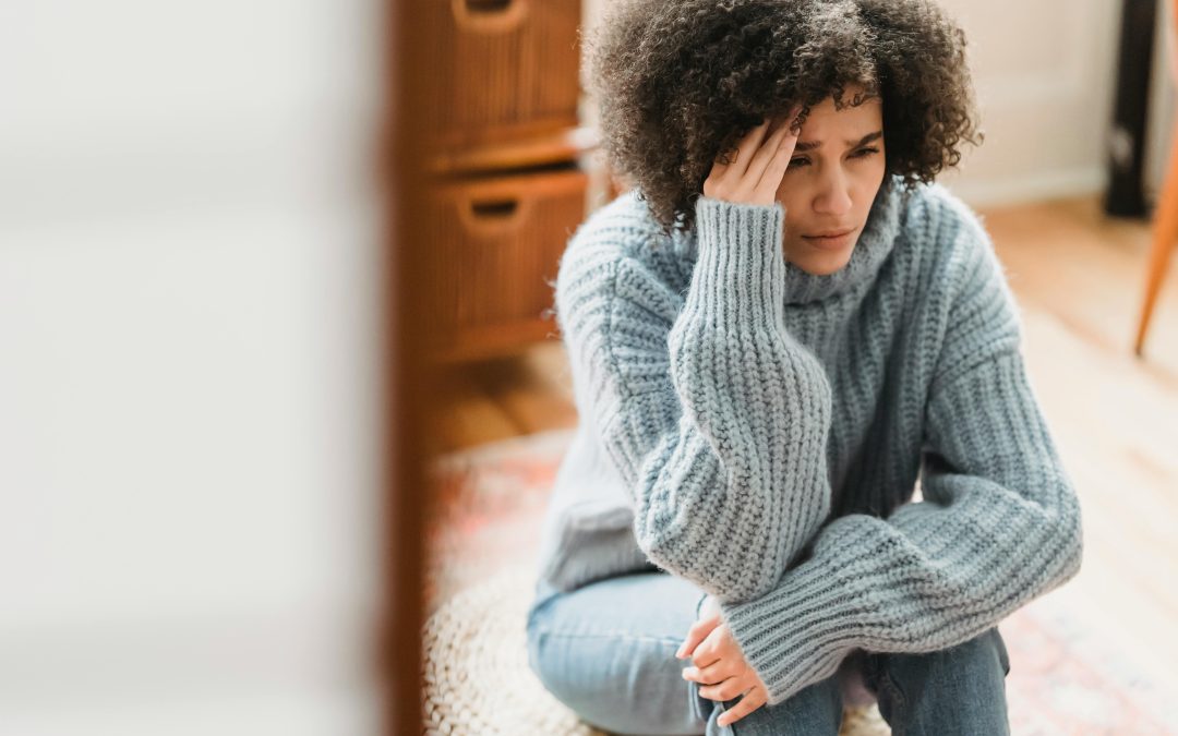 Woman sits on a cushion on the floor with hand at the side of her head looking confused and wondering if she is being emotionally abused by her partner.